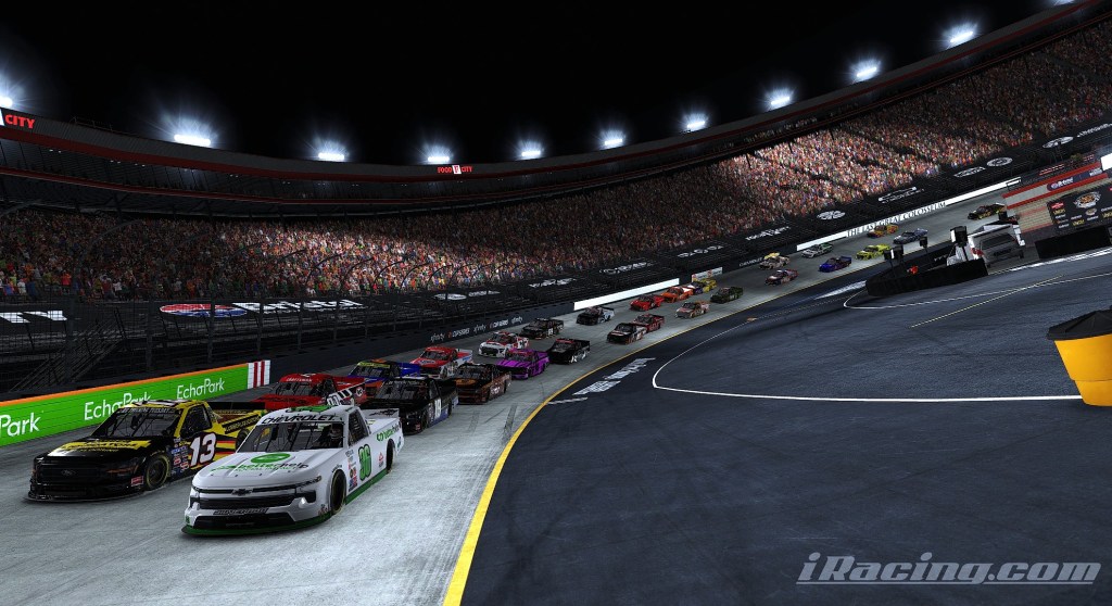 A group of race cars, led by a white vehicle, compete on a well-lit oval track with a cheering crowd in the stands during a nighttime race.