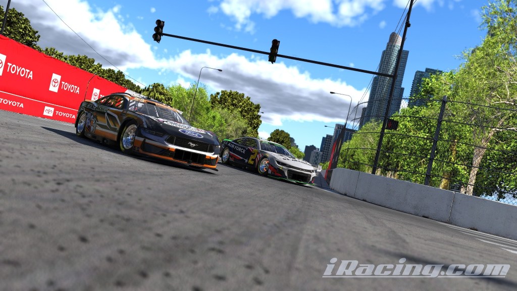 Two race cars navigating a turn on a city street circuit, with a red advertising banner in the background and skyscrapers visible in the distance.