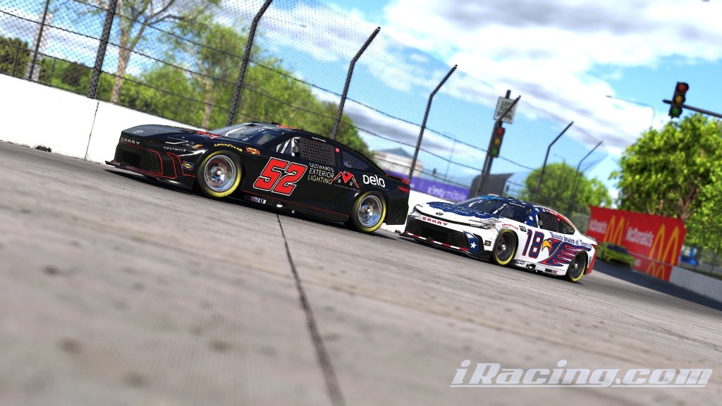 Two racing cars, one black with number 52 and the other white with number 18, are competing closely on a racetrack.