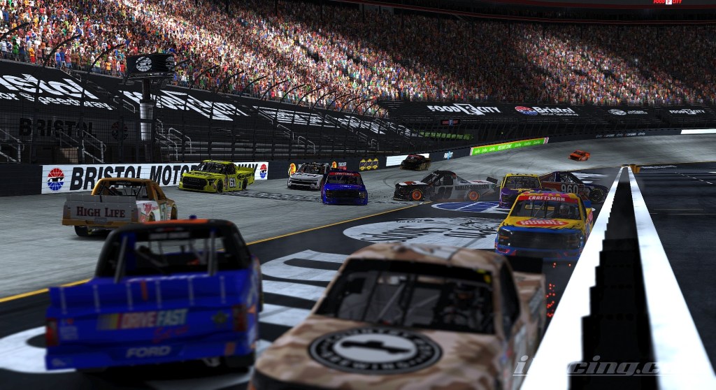 A busy scene at Bristol Motor Speedway during a truck race, with multiple vehicles racing close together under stadium lights and a large crowd in the stands.