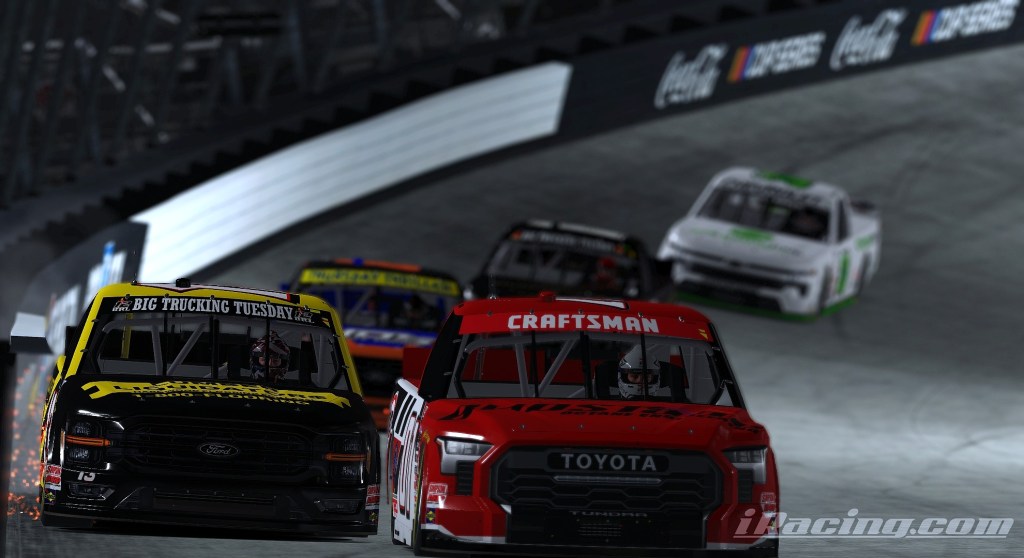 A group of racing trucks competes closely on a banked track at night, with a focus on a yellow truck leading the pack and a red truck closely following.